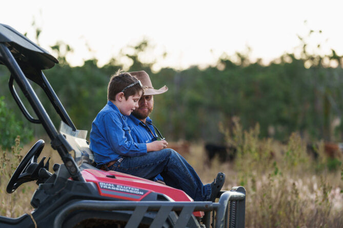 A child sits on the hood of a quad bike on a property and his father looks at something over his shoulder..